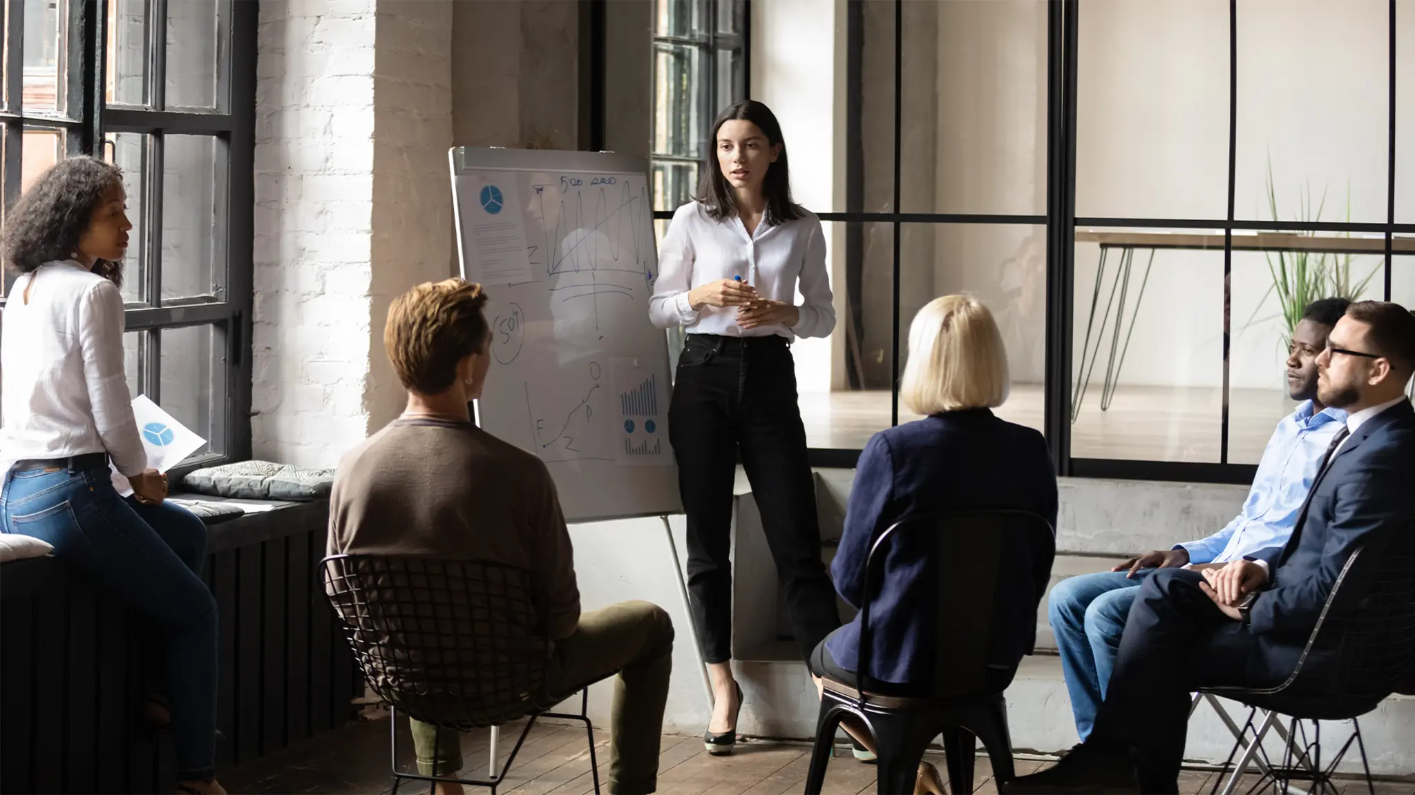 A group of business professionals sitting and listening to a standing presenter next to a flip chart in a well-lit office space with large windows
