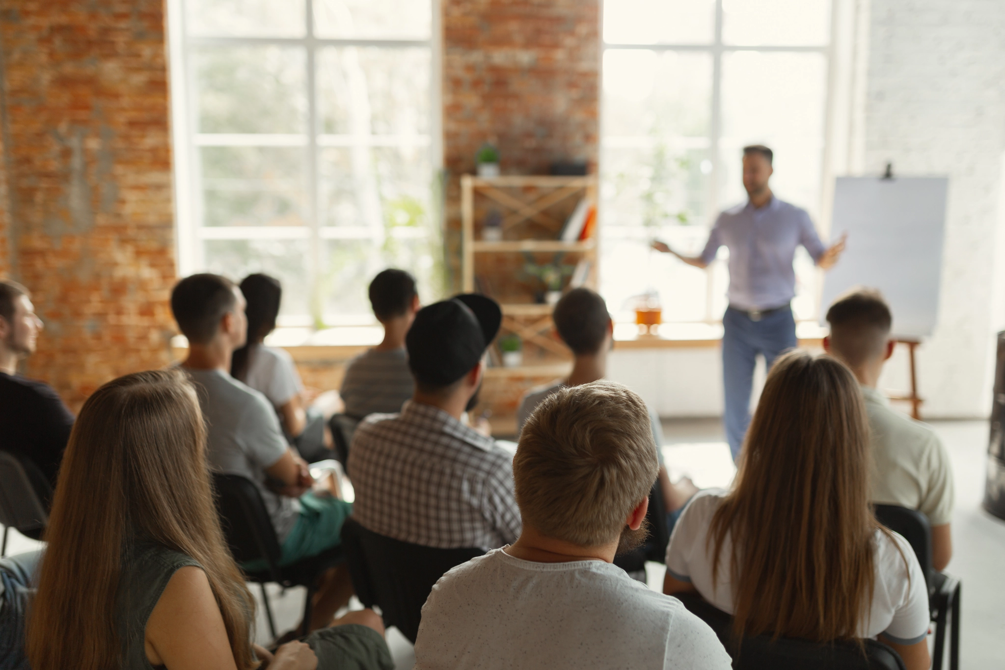 A group of people sitting and listening to a presenter in a workshop or training session in a room with brick walls and large windows