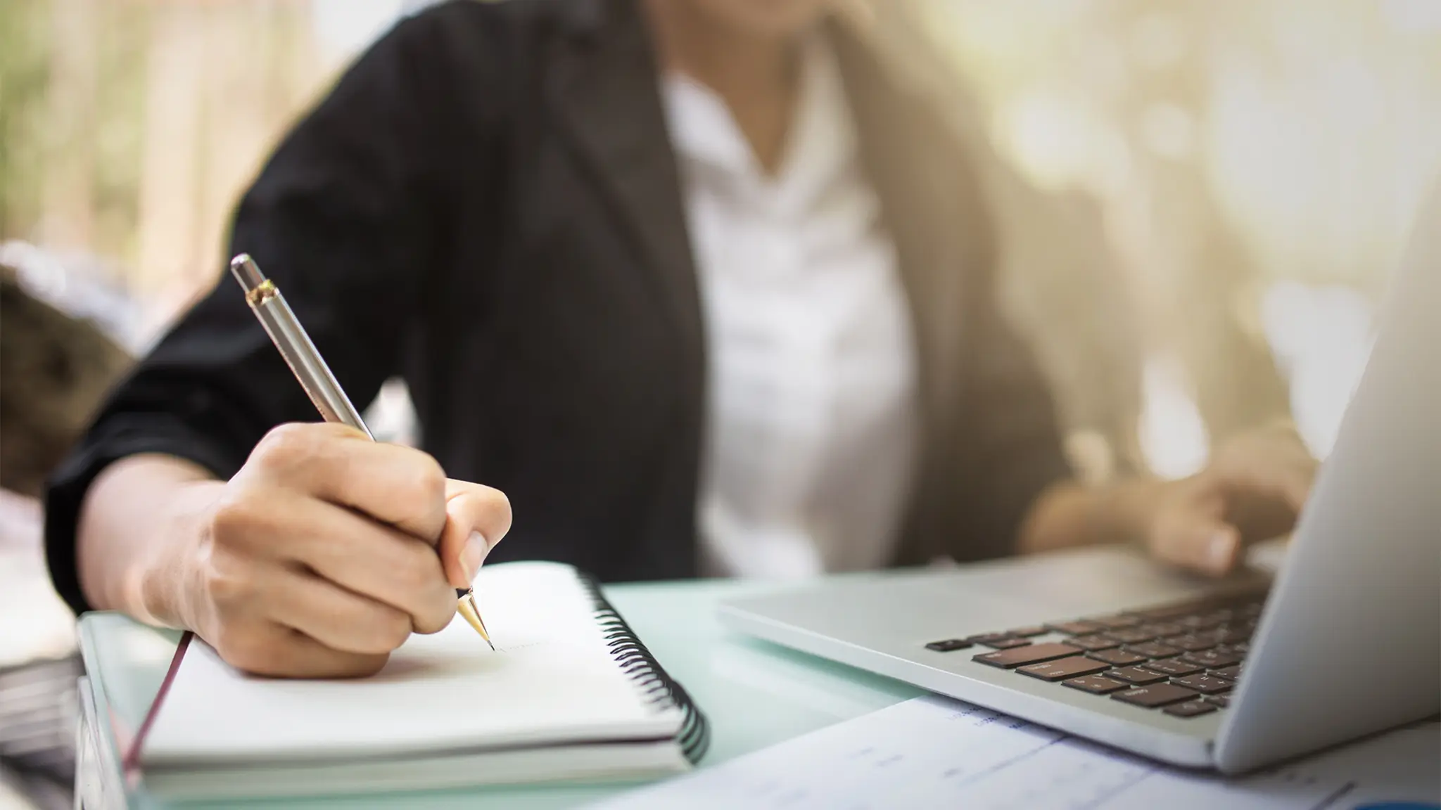 A DBA student at a table uses a laptop while jotting down notes in a notebook, showcasing her concentration and creativity